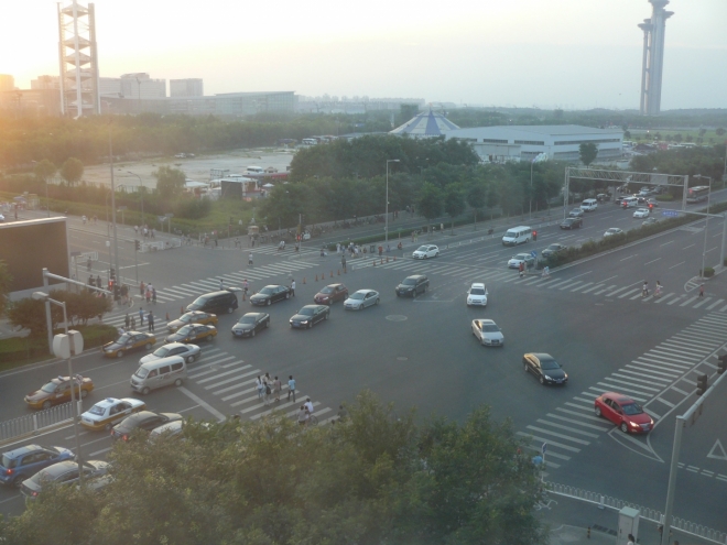 Křižovatka pod hotelem trochu ilustruje divokost tamní dopravy. Na přechodu už přecházejí lidé, přesto proud aut stále ještě dojíždí. V pozadí vlevo je rozhledna v olympijském areálu.