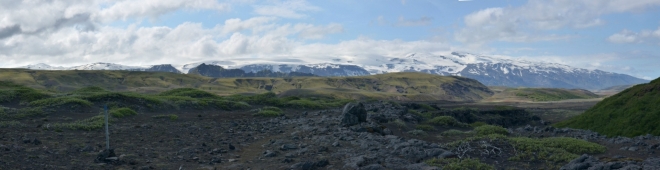 Panorama Eyjafjallajökull. Dole černý písek, v němž se boří naše nohy. Z písku vyčnívají zeleně porostlé kopečky.